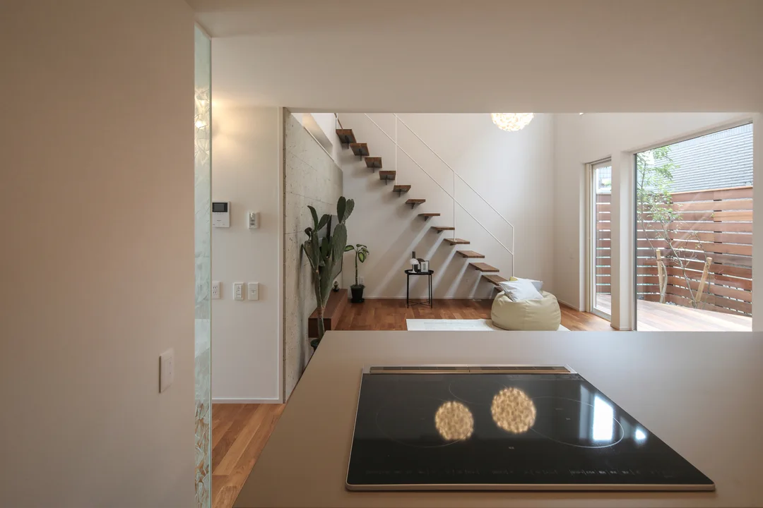 キッチンのIHコンロの前に立って正面を見ている写真。正面には吹抜け階段のあるリビングが見える。