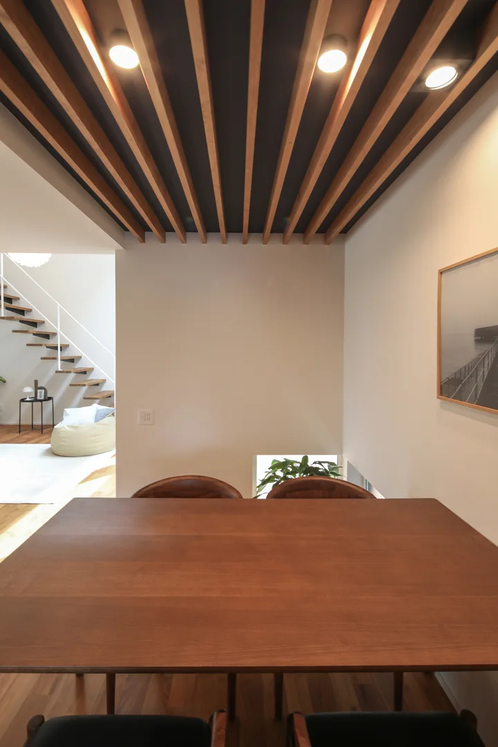 ダイニングスペース。天井は2.1ｍで低めにして落ち着けるようにした。ルーバー天井にして、ルーバーの間からダウンライトでダイニングテーブルの上を照らしている。右手の壁にはアート写真が掛かっている。