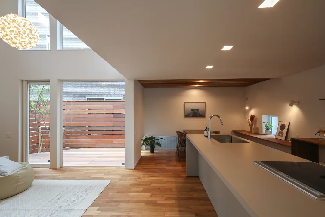 リビング、ダイニング、キッチン。リビングの向こうにはアウトリビングが見える。アウトリビングはウッドデッキの床で、隣地との目隠しにウッドフェンスを設けている。リビング上部のみ吹抜けで、ダイニングキッチンの天井は2.1ｍと低く設定している。リビングの大きな丸いペンダントライトが際立っている。