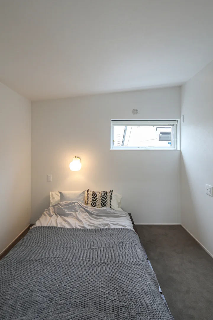 主寝室。床はグレーのタイルカーペット。セミダブルのベッドが置かれていて、枕もとの壁からはルイスポールセンのウォールライトが出ている。壁と天井は白いクロスで、落ち着いた生活感のある空間になっている。