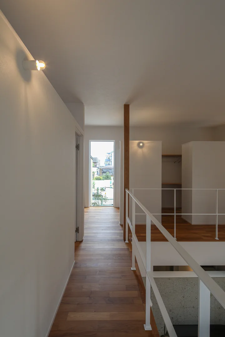 2階廊下。正面には縦長窓があり、そこから光が入ってくる。右手は吹抜け部分で、白いアイアンの手すりがL字に走っている。照明は壁付けのシンプルなもの。チークの無垢フローリングに、白い壁と天井。