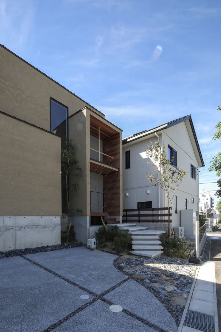 家の外壁は、明るすぎないベージュ。四角い3つの箱を組み合わせたようなデザイン。ベージュの塗り壁はジョリパットを使用しています。中央の玄関に向かってコンクリートの板が浮いたような階段があり、その先はウッドデッキの玄関ポーチになっています。ポーチの上部には庇が出ていて、庇の上には十字の飾りがついています。