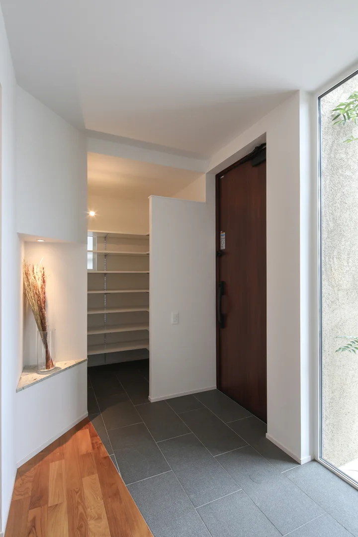 2.5帖の玄関ホール。壁紙は白、床には、硬くて水にも強い、御影石を使用しました。室内側の床は世界三大銘木の1つ、チークの無垢フローリングです。正面にはシューズクロークがあり、袖壁で目隠しになっています。向かって右手には床から天井までの縦長窓を設けました。