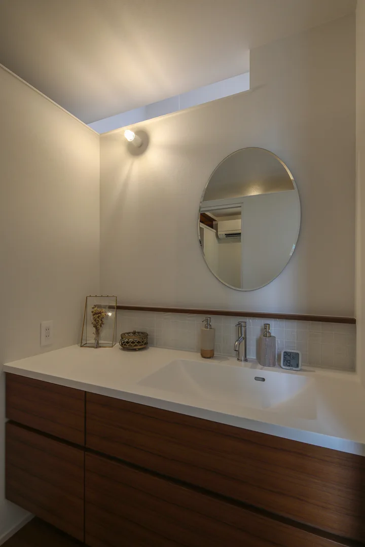 造作洗面台。白い人工大理石の天板と、床と同じチーク材の扉。オーバルの鏡を添えることで、固くなりすぎないゆるやかな雰囲気に仕上げています。家具のような洗面台が主役ですので、照明はシンプルな壁付けにしました。
