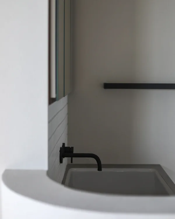 造作洗面台。水栓は壁だしのマットなブラック。シンクは白の病院シンク、天板はグレー。腰壁は一部アールがついている