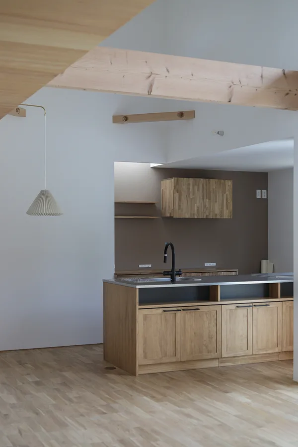 オープンなキッチンを見た眺め。右側にウッドワンのパーティータイプのシステムキッチン、左側にはダイニング用のペンダントライトが下がっている。床は明るいオークの無垢フローリングで、壁紙は白、キッチンの後ろだけココアのような茶色のアクセントクロスが貼られている。上部には梁が見えており、吹抜になっていることが分かる