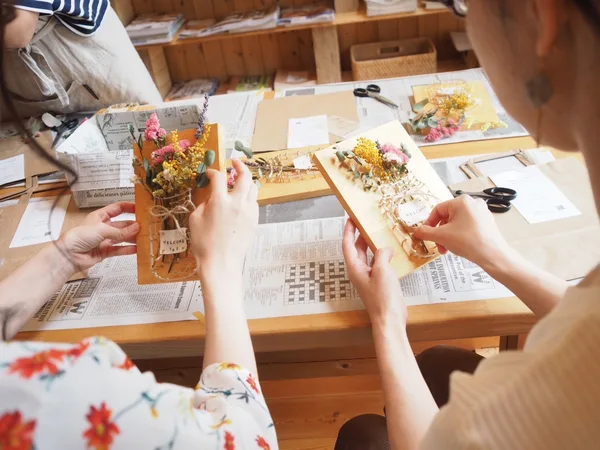 大人の片手サイズの四角い板に釘を打ち、そこに糸が掛かっている。糸はジャムの瓶のような画を描いており、ドライフラワーがその瓶に刺さっている。それらを2人の女性が互いに手に持って見せ合いながら語らっている