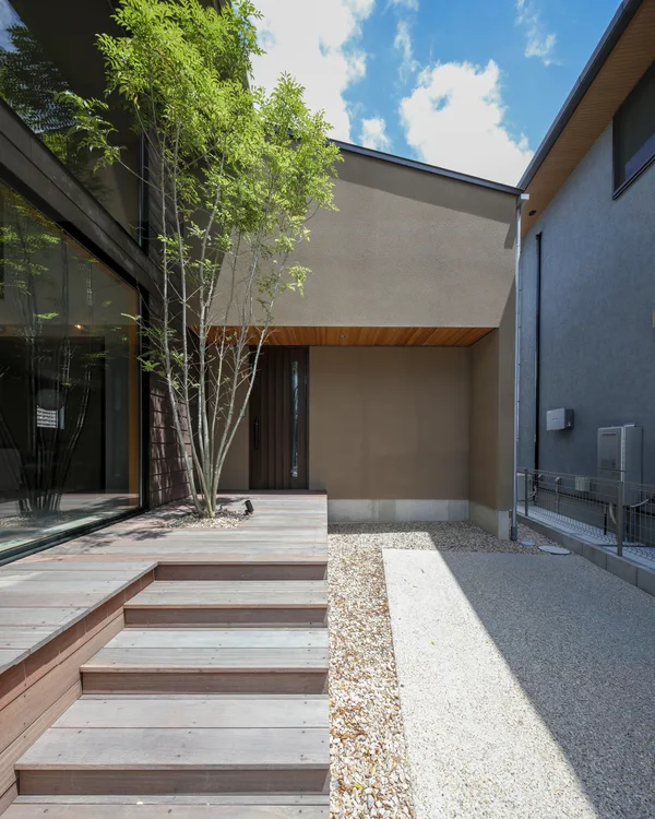 手前にウッドデッキと階段、突き当りにはグレーの壁の家。茶色の玄関ドアがある。左側には緑のシマトネリコ。