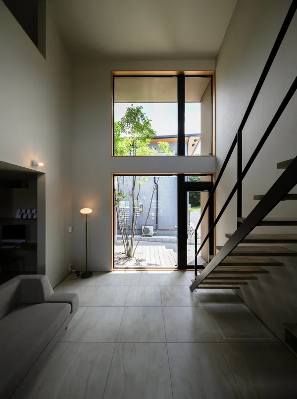 室内から窓の外を見ている。壁はグレージュのクロス、床は壁の色に合わせた60cm角のタイル張り。吹き抜けていて、吹抜一面に4枚の窓がある。うち1枚はテラスドアになっている。窓の向こうにはシマトネリコの緑と隣家、青い空が見える。右手にはスケルトン階段があり、踏板の裏側が見えている。