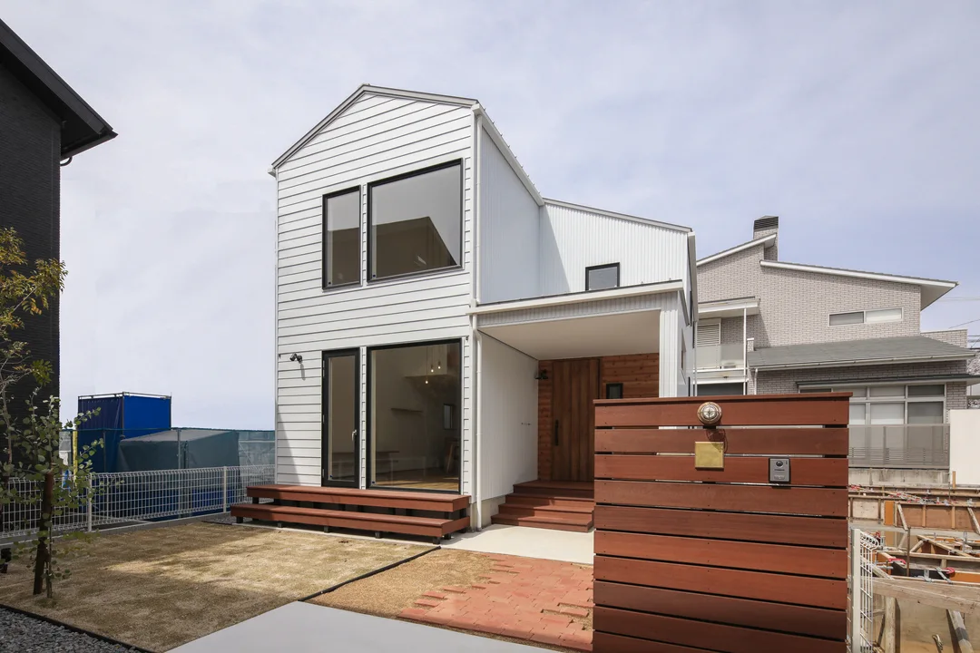 三角屋根の白い家を向かって右斜め前から見た写真。右手前にはセランガンバツ製の門塀。門塀には照明とインターホン、表札がついている。家の右側は深い軒。軒の奥は木板張りと玄関ドア。家の左側には田の字の窓。家の前、向かって左手に芝生の庭