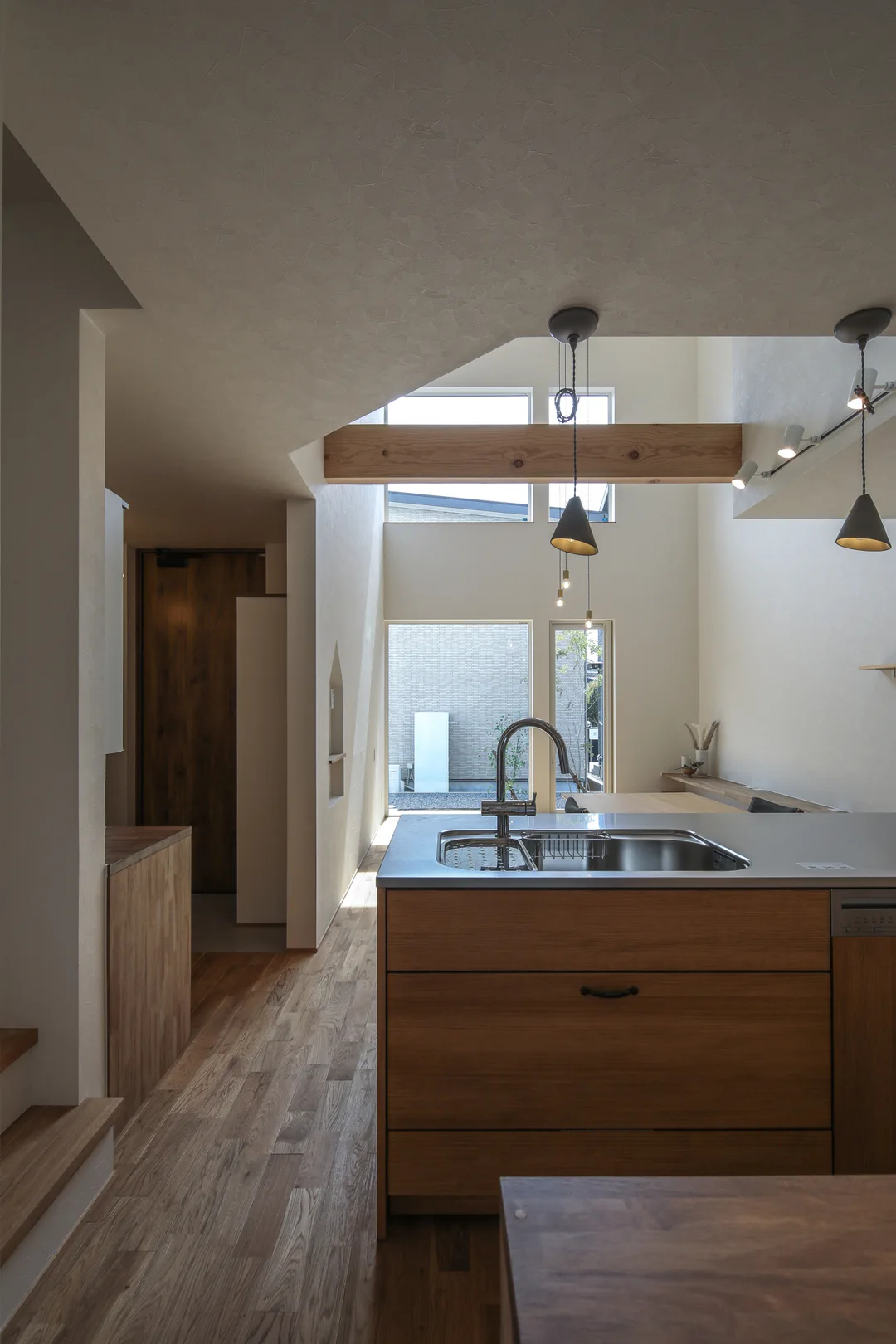 手前にキッチン、奥に窓が見える。キッチン上部にはペンダントライトが2灯見える。無垢フローリングの床と白いクロス。