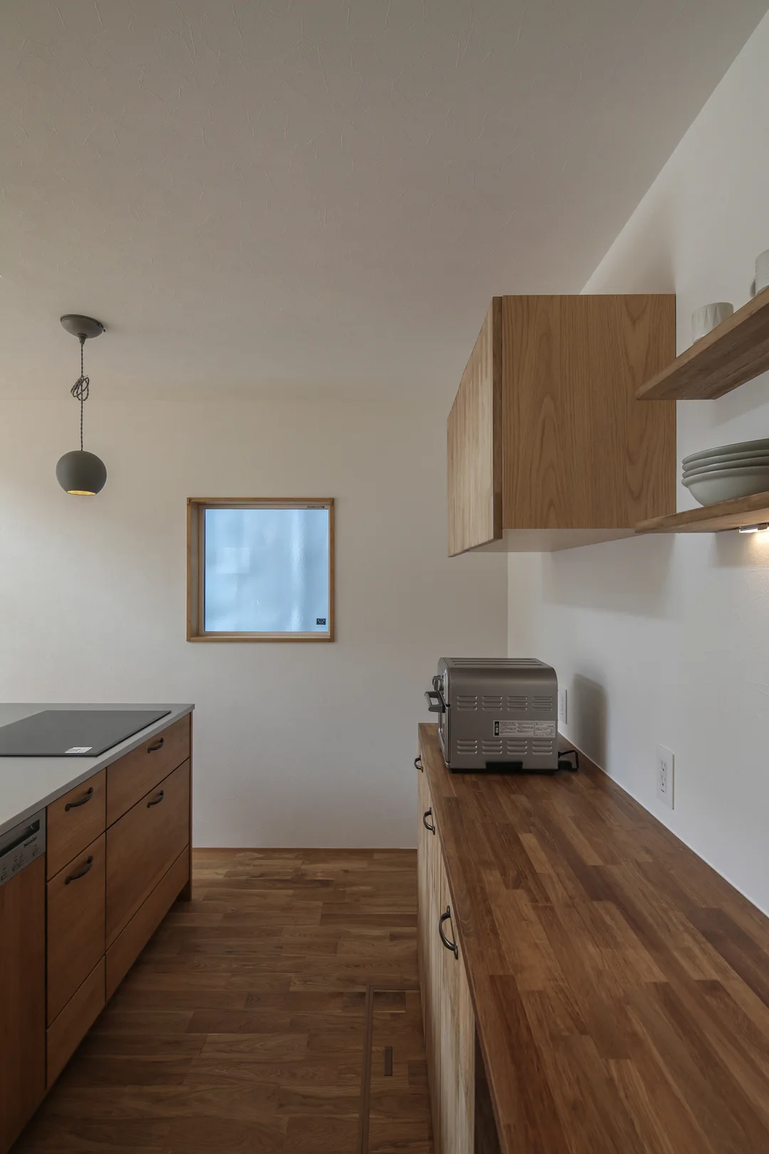 右手にカップボード、右手上部に飾り棚と吊戸棚。左側にキッチン。奥に四角い窓。