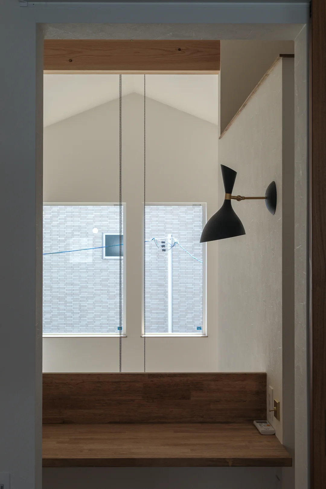 右手の壁に黒いシェードのウォールライト。下に木の机。正面には吹抜けの窓が見える