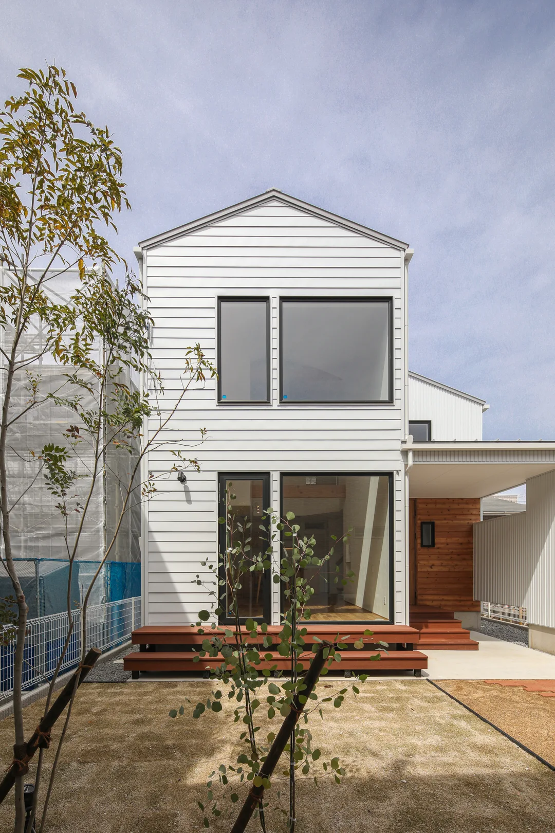 縦に長い三角屋根の白い家。田の字型に窓がある。窓枠は黒。手前に植物が2本。奥は青空に薄雲