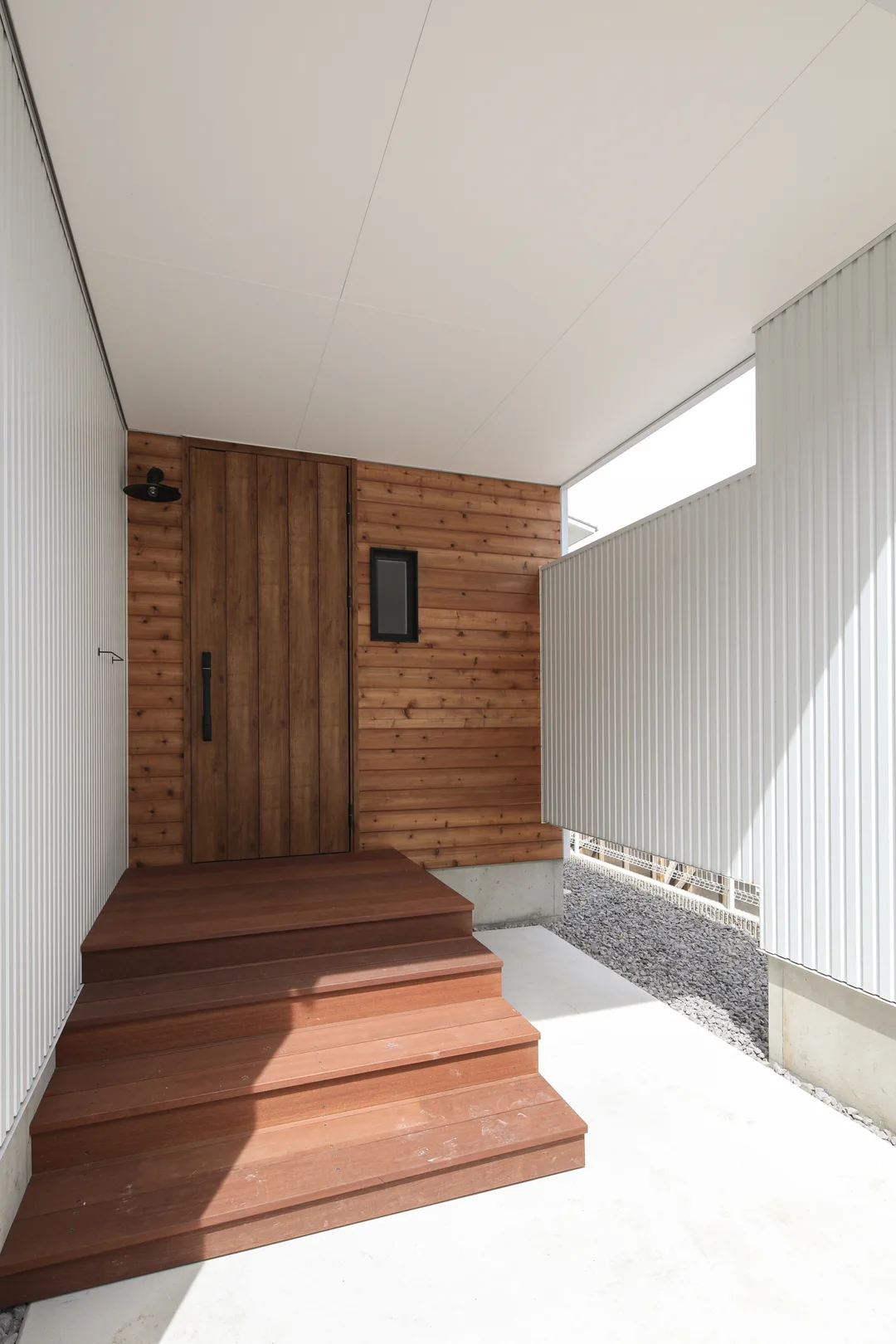 庇と袖壁がある凹んだ空間。突き当りの壁は木板張り。木目調の玄関ドアがある。ウッドデッキ製の玄関前の階段