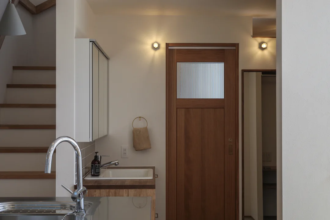 室内。正面右手に木製のドア。ドアには模様のあるガラス窓がある。ドアの上部両側に壁付け照明。左手前にキッチンの水栓。キッチンの奥に2階への階段がある