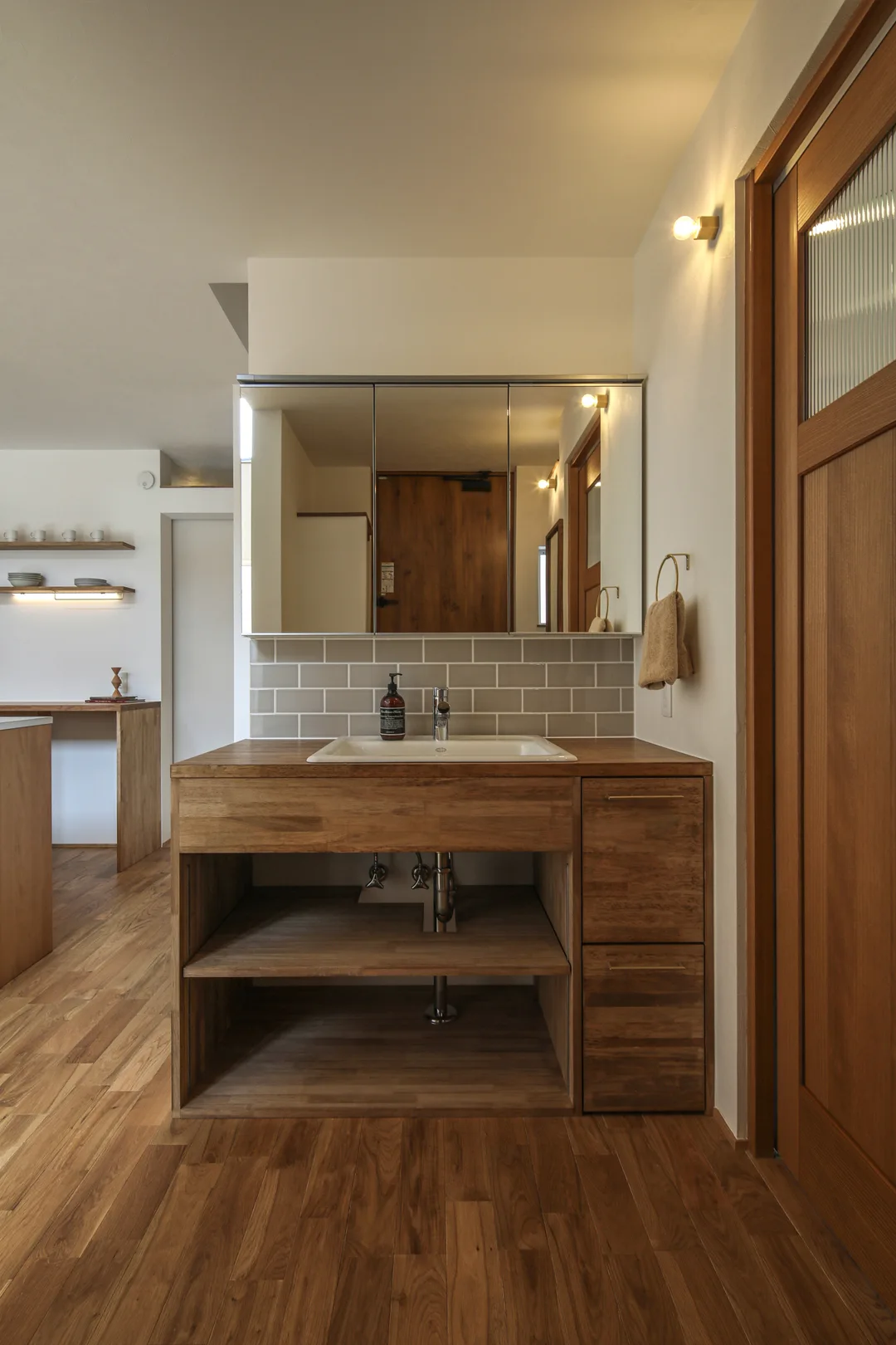 洗面台。キャビネットは濃い茶色で木製。洗面器は白い琺瑯製でキャビネットに埋め込み。水栓はクローム。鏡は横長四角の三面鏡。三面鏡とキャビネット天板の間の壁にグレーのタイルが馬目地で貼られている。