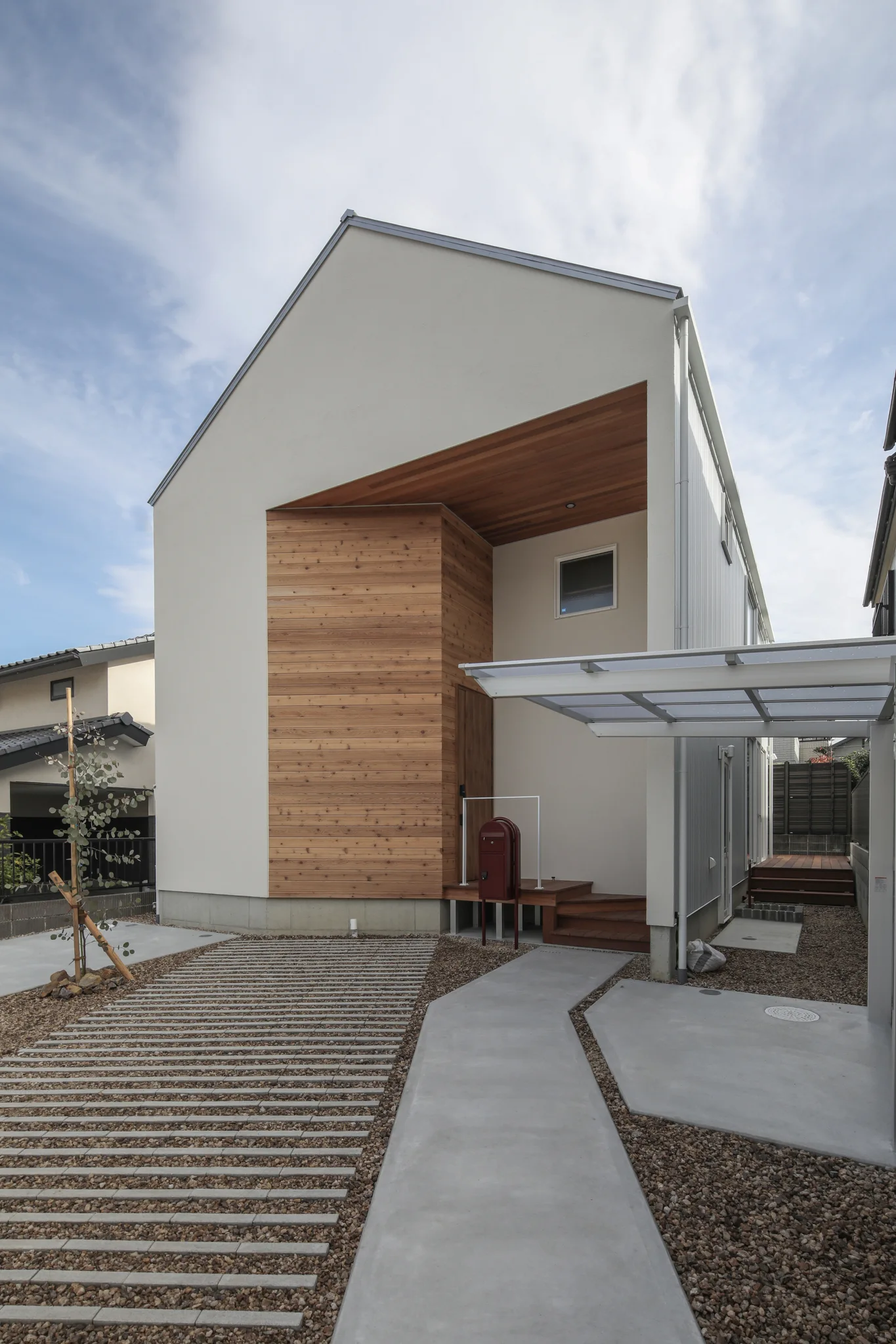 三角屋根の白い家を向かって右斜め前から見た写真。三角屋根の下に凹みがあり、その部分が玄関ポーチになっている。玄関ポーチまでは土間コンクリートの道がある。向かって右手にはサイクルポートもある。