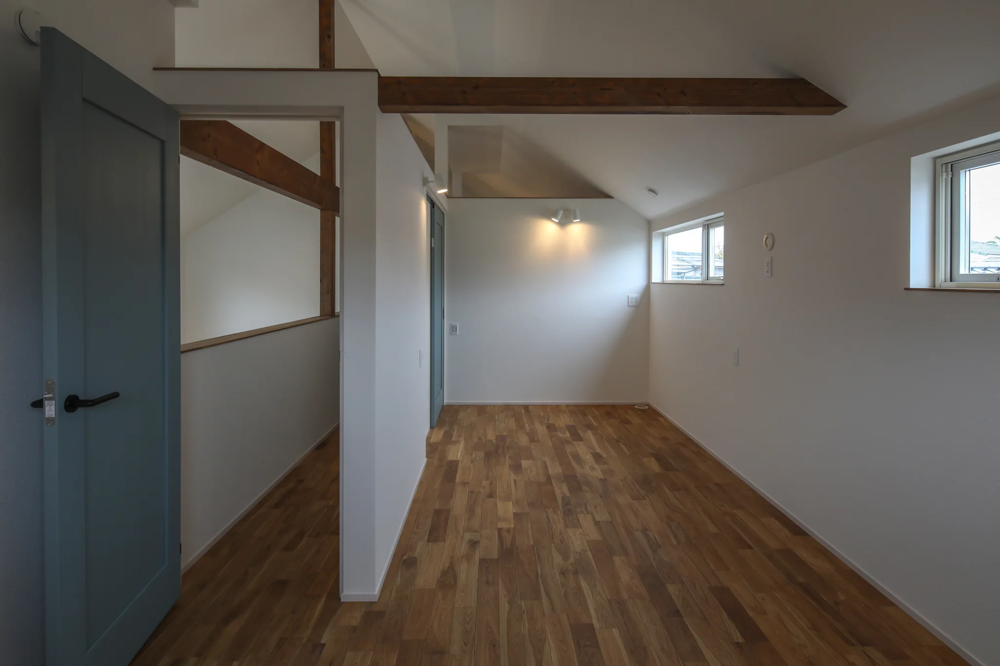 子供室。2部屋の間には壁が無く、将来壁を立てるための梁が通っている。左側にはブルーグリーンの引き戸がある。照明は壁付けのスポットライト。窓が2つあいている。