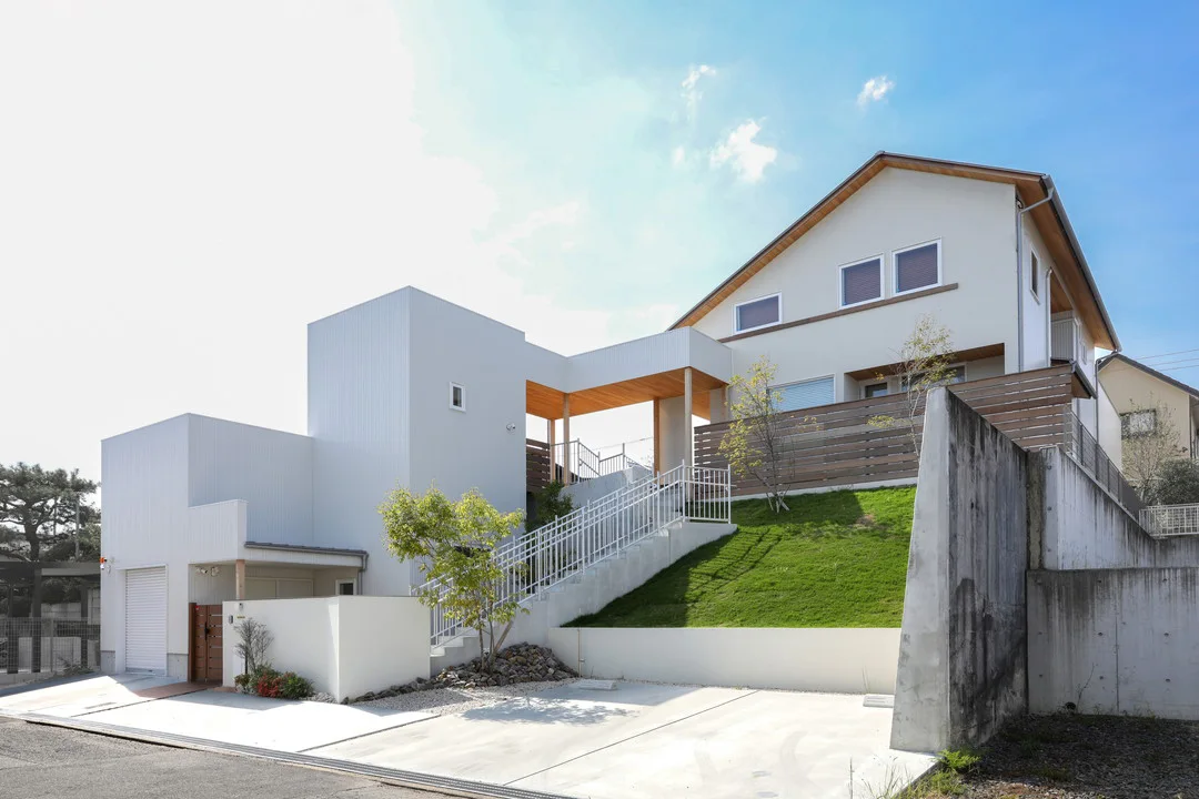青空の屋外。手前に擁壁がある。奥には白い箱型の建物と三角屋根の家がある。家には横に3つ並んだ窓がある。所々に木が植わっている。家から道路までは高低差があり、法面には芝生や植物が植えられている。