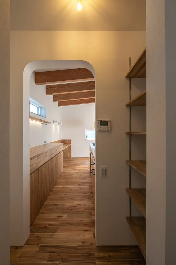 木の床、木製のカップボードとカウンター、アーチ型の入口がある、モダンなデザインのキッチンです。露出した梁が天井にあり、部屋の奥には窓があります。