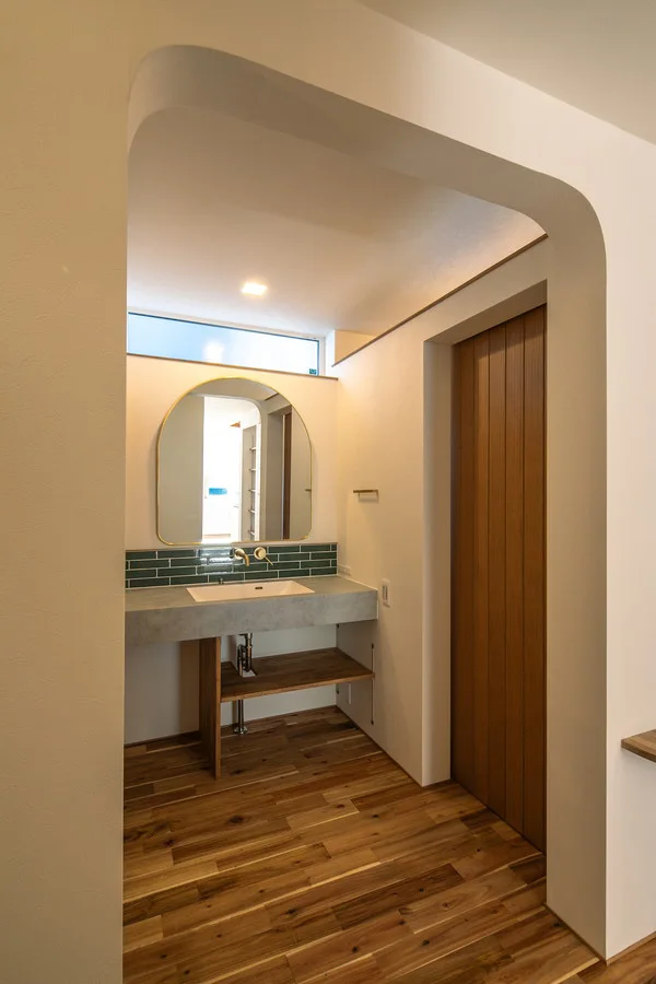 木の床とアーチ型の入口がある、モダンなデザインの洗面所です。コンクリートのように見えるカウンター、緑色のタイル張りのバックスプラッシュ、丸い鏡があります。隣には木製のドアがあり、鏡の上には横長の窓があります。