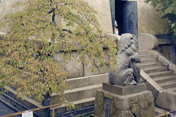 祇園閣の入り口部分。手前に木が植わっていて黄色い石垣が見える。狛犬のように座っているのはライオンの石像。