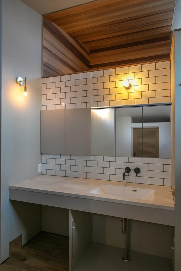 洗面。グレーの扉付き収納と鏡が付いている。水栓は壁付で黒色。奥にはレッドシダーの羽目板張りが見える。