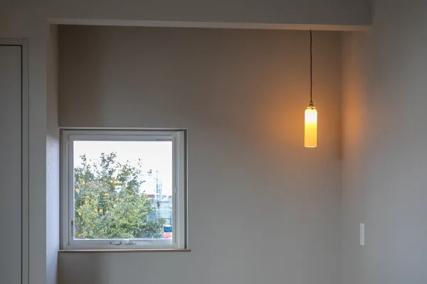 グレーの壁紙の部屋。四角い窓とペンダントライトがある。窓の向こうには木が見える。ライトの光はオレンジ色をしている。