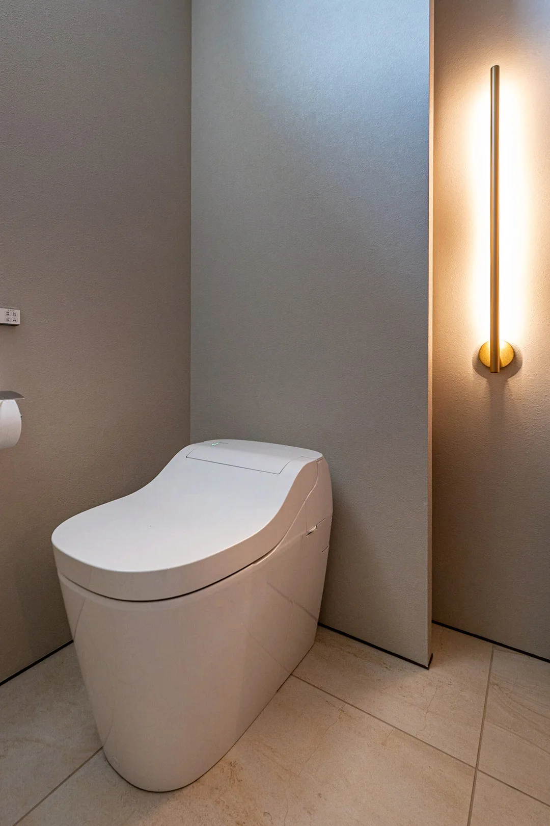 モダンなトイレ。シンプルな白いトイレと、少し凹ませた背面の壁に取り付けられた照明器具が見える。床はタイル。