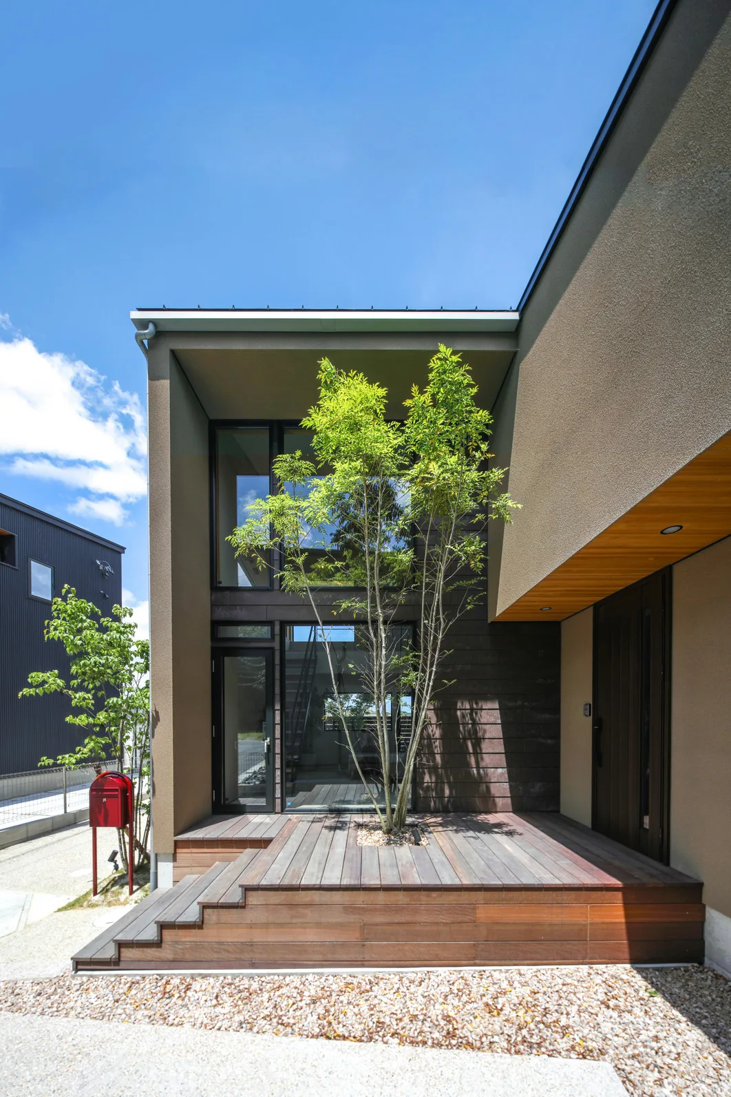 晴れた日に撮影された日本のモダン住宅の正面玄関。木製の階段とデッキ、大きな窓、そして玄関ドアが見える。デッキには穴が開いていて2階の軒まで届く木が植わっている。