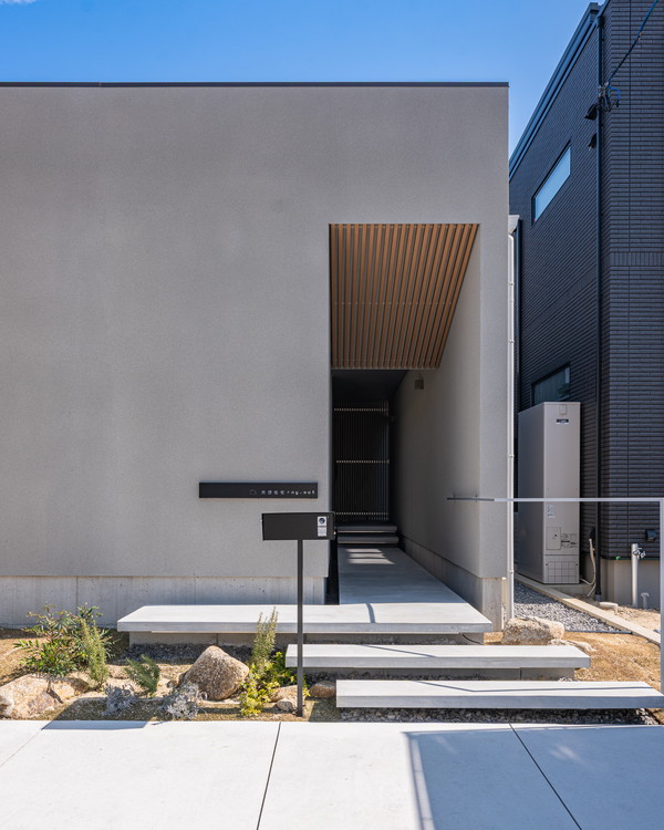 グレーの外壁に四角い穴が開いて言いて奥にコンクリートのアプローチが伸びている。軒天には格子が貼られている。