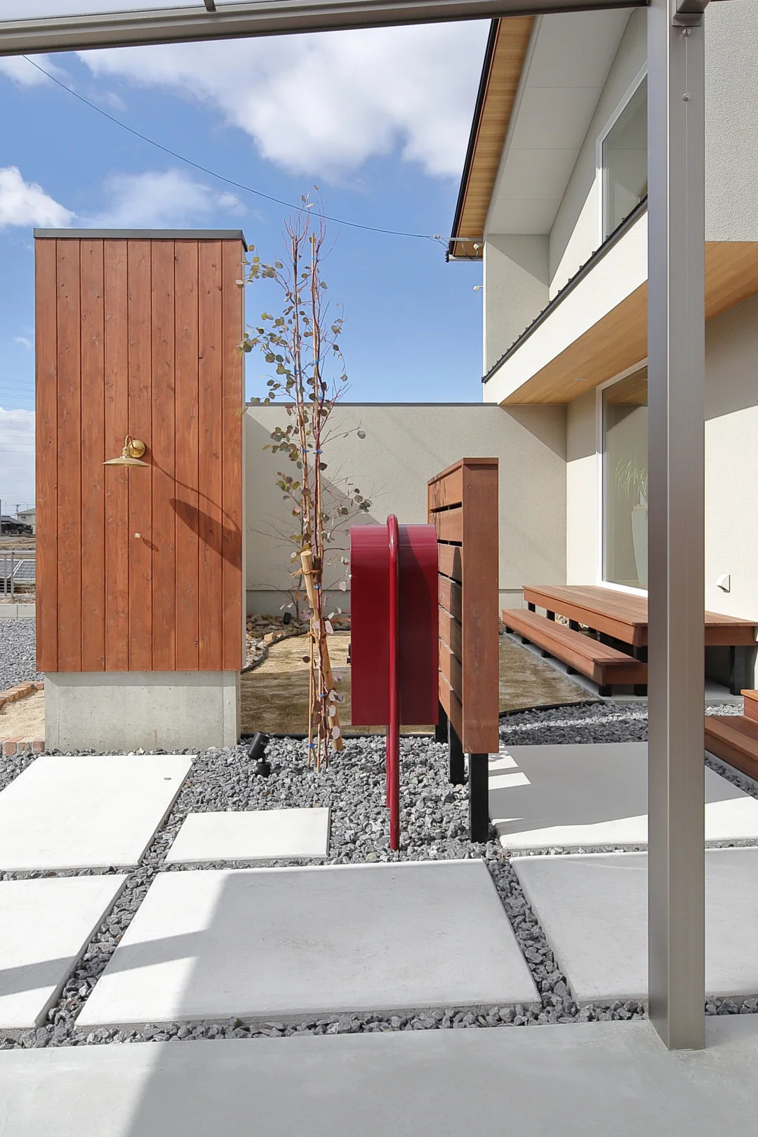 住宅の玄関アプローチ。木製の外壁、赤いポスト、シンボルツリー、コンクリートと砂利でデザインされた床が見える。