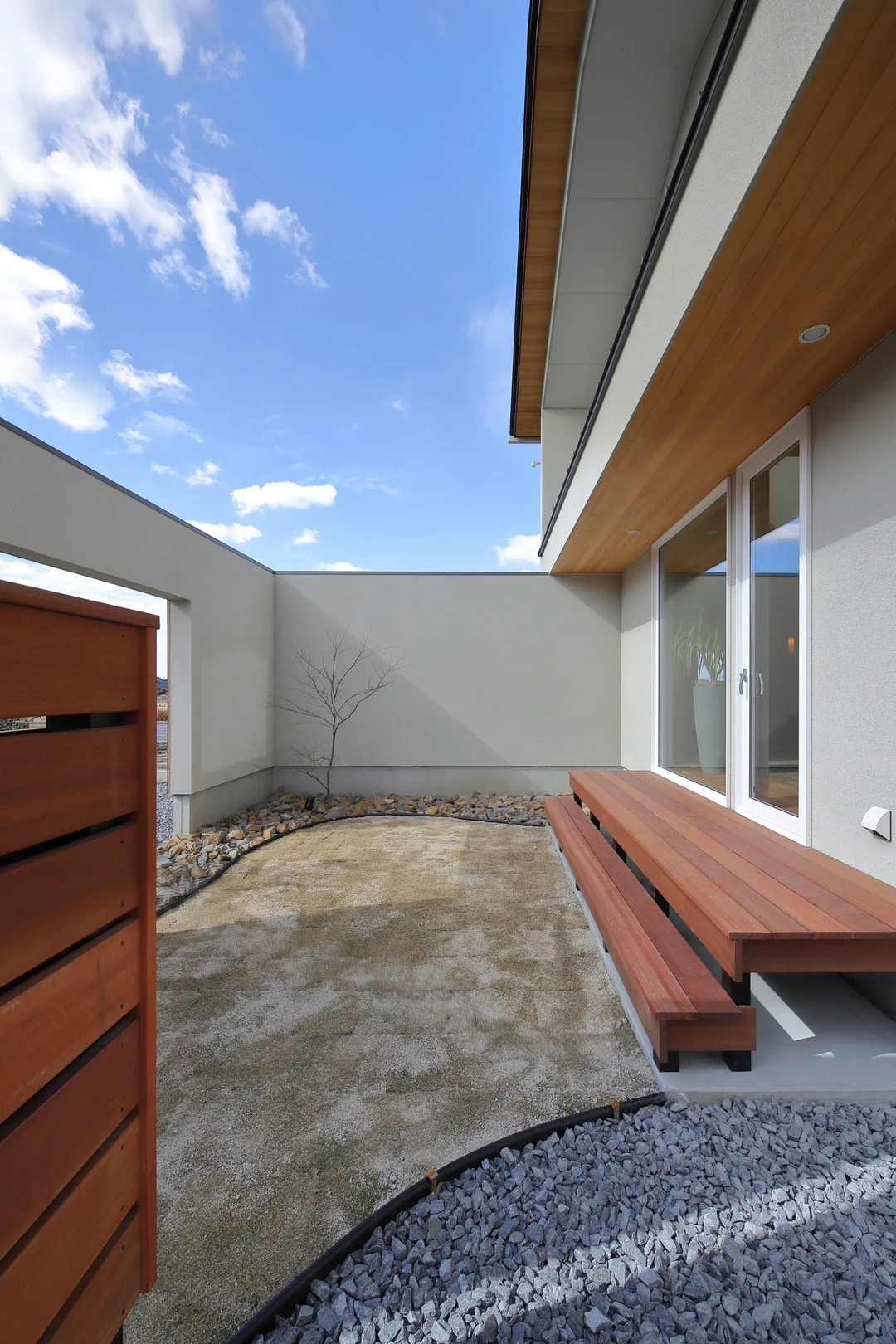 住宅に隣接する中庭。ウッドデッキ、白い壁、砂利が敷かれ、壁際には木が植えられている。