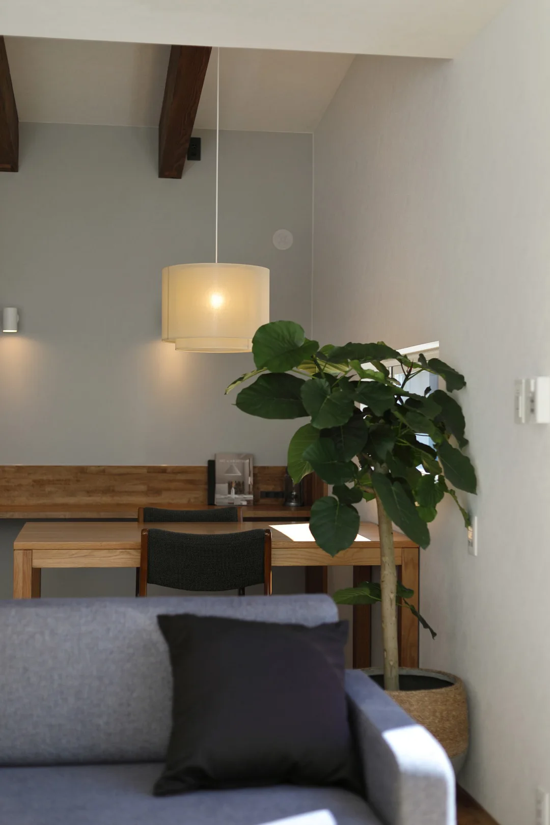 リビングの一角。木製のカウンターデスクと椅子、手前にはグレーのソファと観葉植物、天井からは布シェードのペンダントライトが吊るされている。