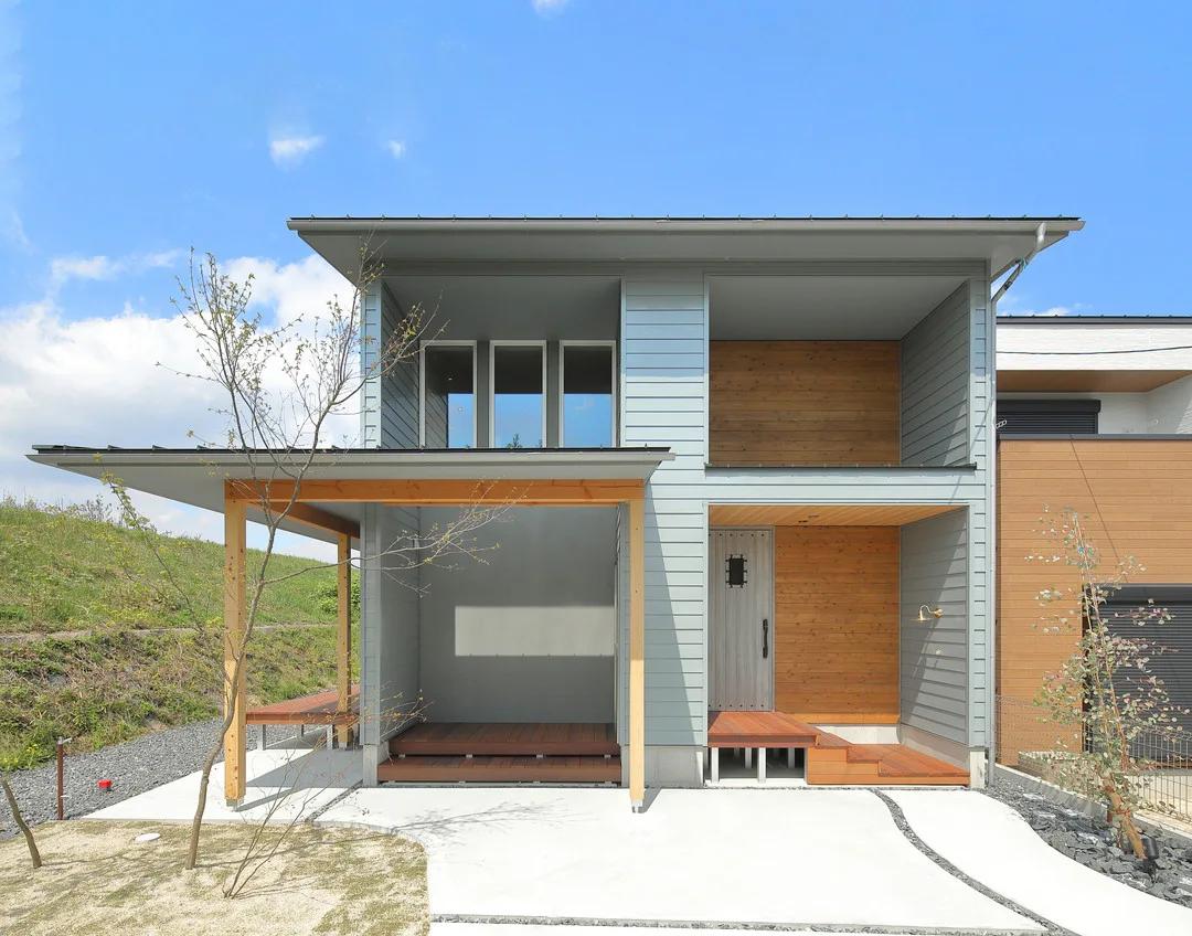 青空の下に建つ、グレーのガルバリウムと木材のアクセントが特徴的なモダンな二階建て住宅。広い屋根付きのポーチとウッドデッキがある。