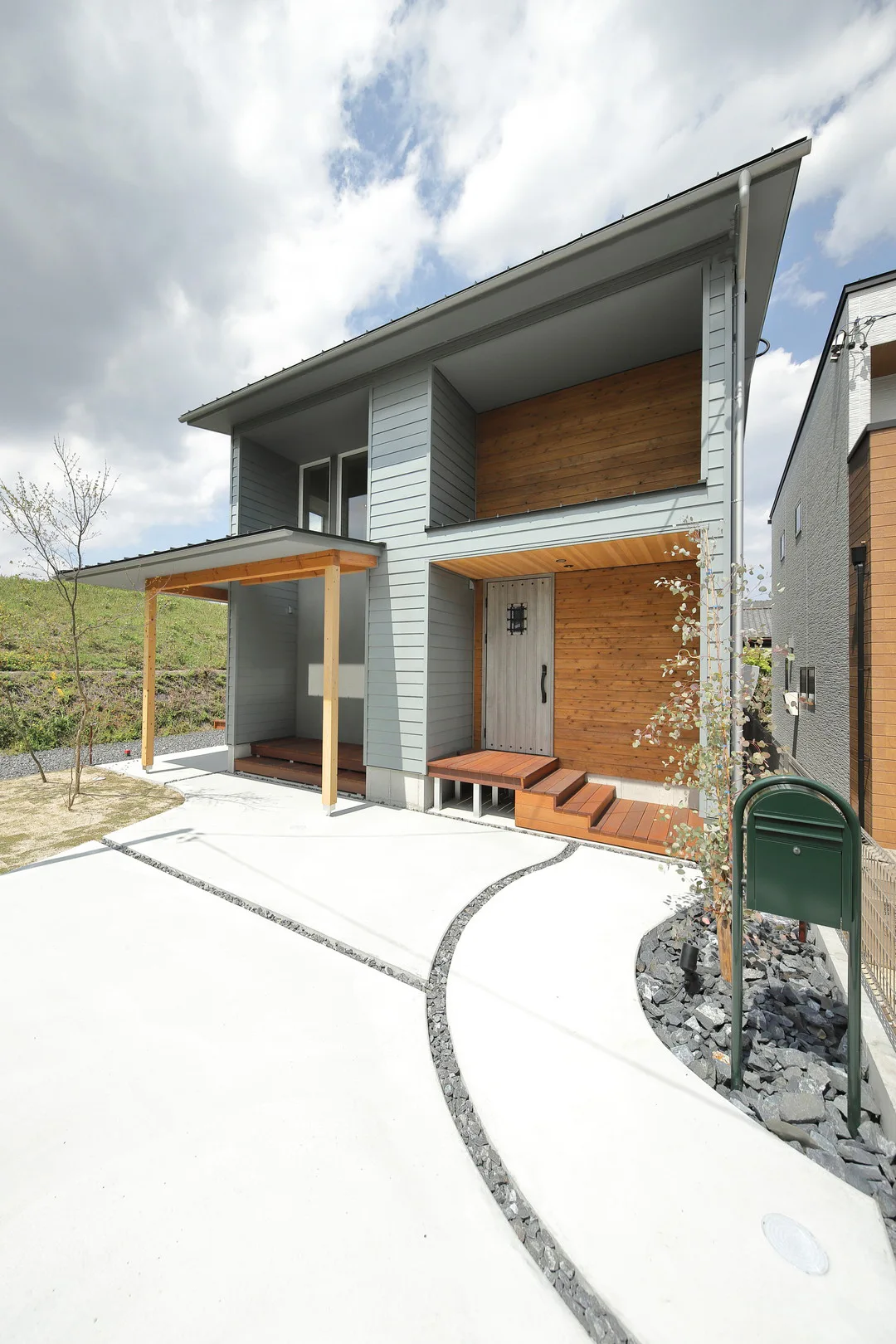 曲線のコンクリート製アプローチがあるモダンな住宅の正面。アプローチの縁は黒い砕石で飾られ、緑色の郵便受けが置かれている。
