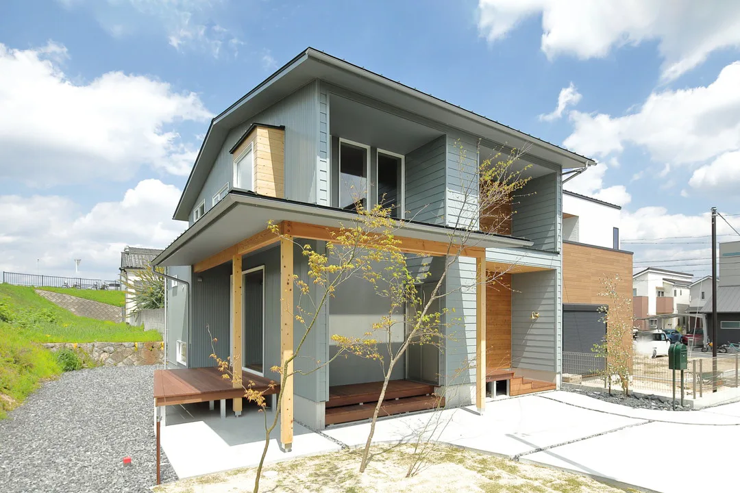 グレーと木を基調としたモダンな二階建て住宅の斜めからの広角写真。屋根付きのポーチや新しく植えられた若木がある庭の様子がわかる。