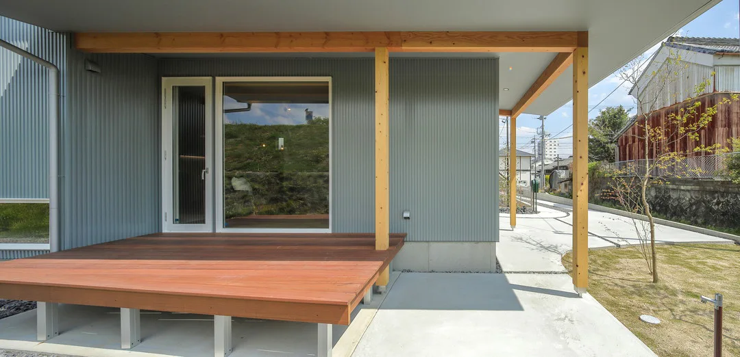 グレーのガルバリウムの外壁を持つ家の側面に沿って続く、屋根付きのウッドデッキ（縁側）。明るい色の木製の柱が屋根を支えている。