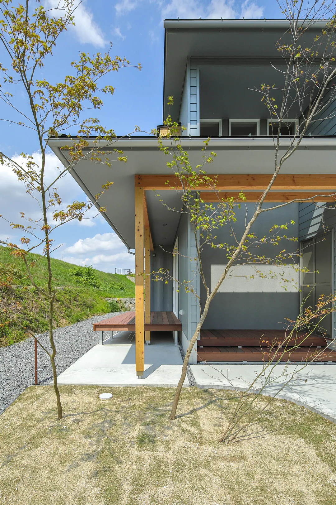 モダンな住宅の側面にある屋根付きのスペース。通路を挟んで二つのウッドデッキが配置され、手前には2本の若木が植えられている。