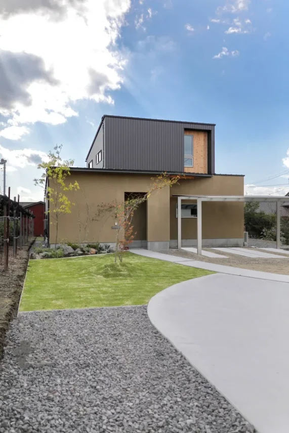青空の下、ベージュとグレーの外壁の家が建っている。手前には緑の芝生と植栽、土間コンクリート、カーポートがある
