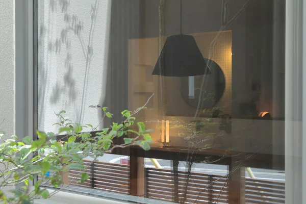 窓辺の景色。手前には緑の葉。窓の奥には黒いペンダントライトと、丸い鏡がある
