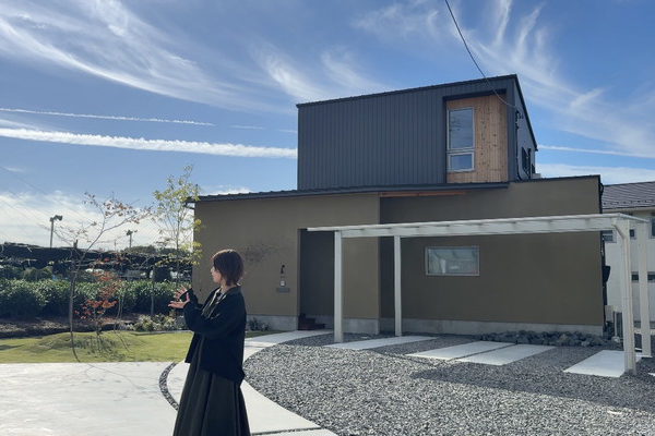 青空を背景に、茶畑と家が並び、手前に黒い服を着た人が立っている