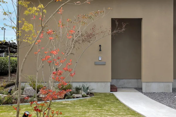 一戸建ての家のファサード。長方形の開口部があり、その手前に植栽がある。開口部に向かってコンクリートの道があり、道の左側には芝生がある