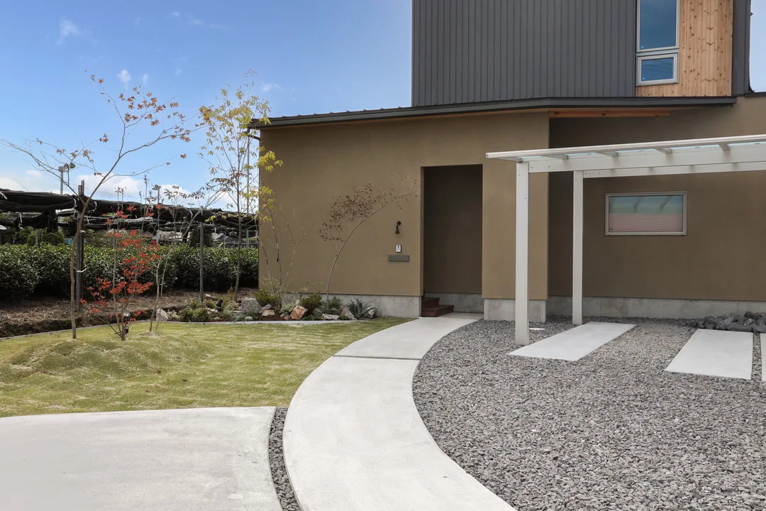 青空の下、ベージュと黒のツートンカラーがモダンな外観の2階建て住宅と、手入れされた前庭