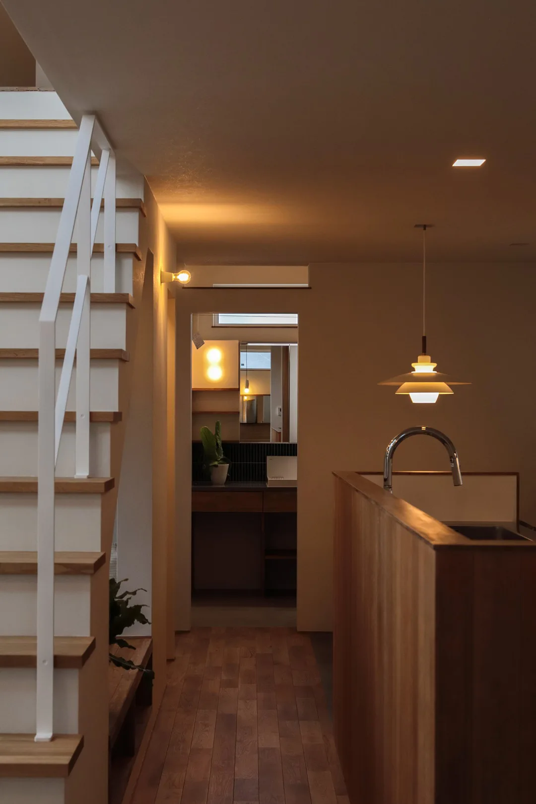 木目の腰壁があるキッチン越しに見る廊下と階段。ペンダントライトや奥の洗面所の灯りが温かみを感じさせる
