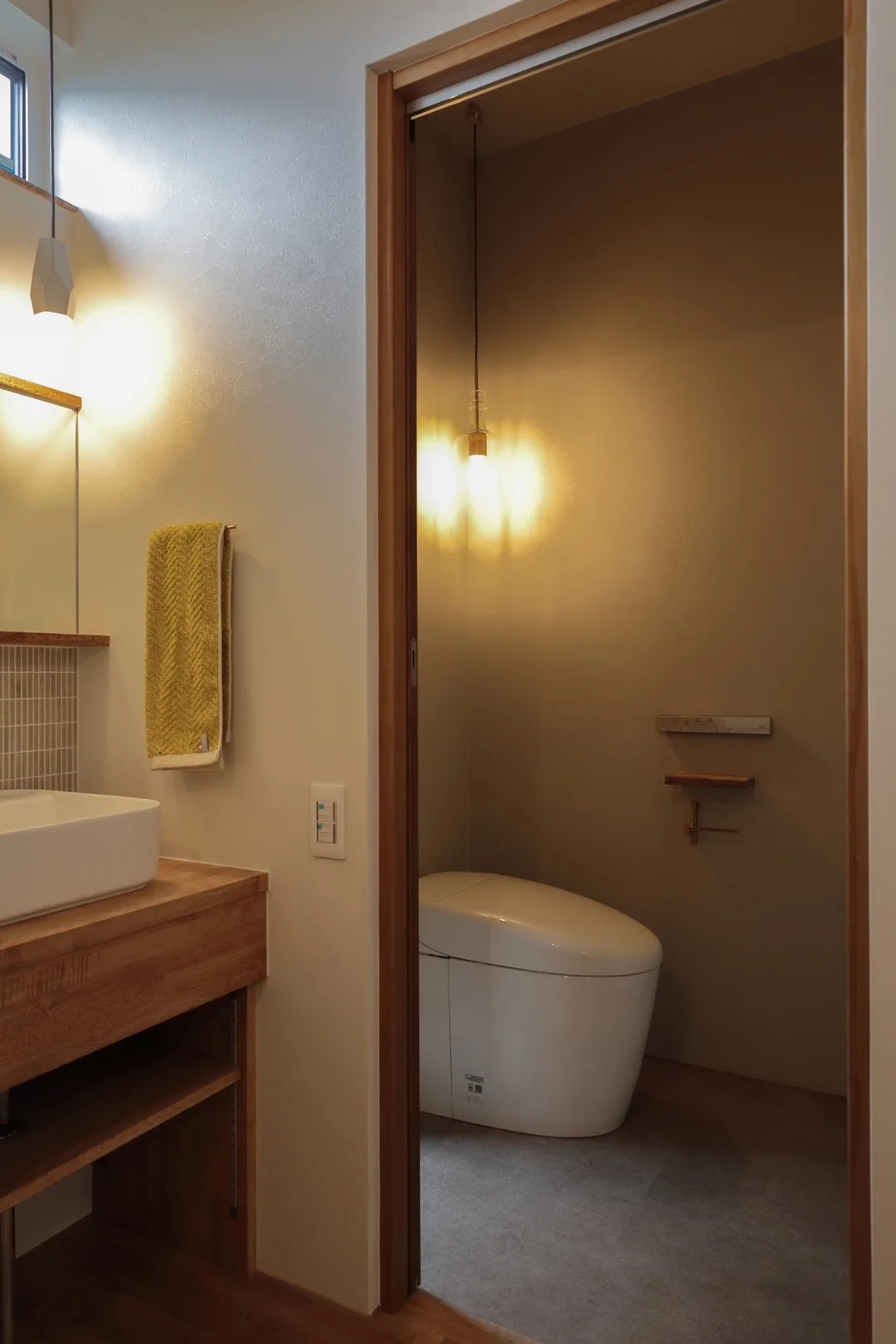 洗面所の隣にあるトイレ。引き戸が開いており、白いタンクレス便器とペンダントライトが見える