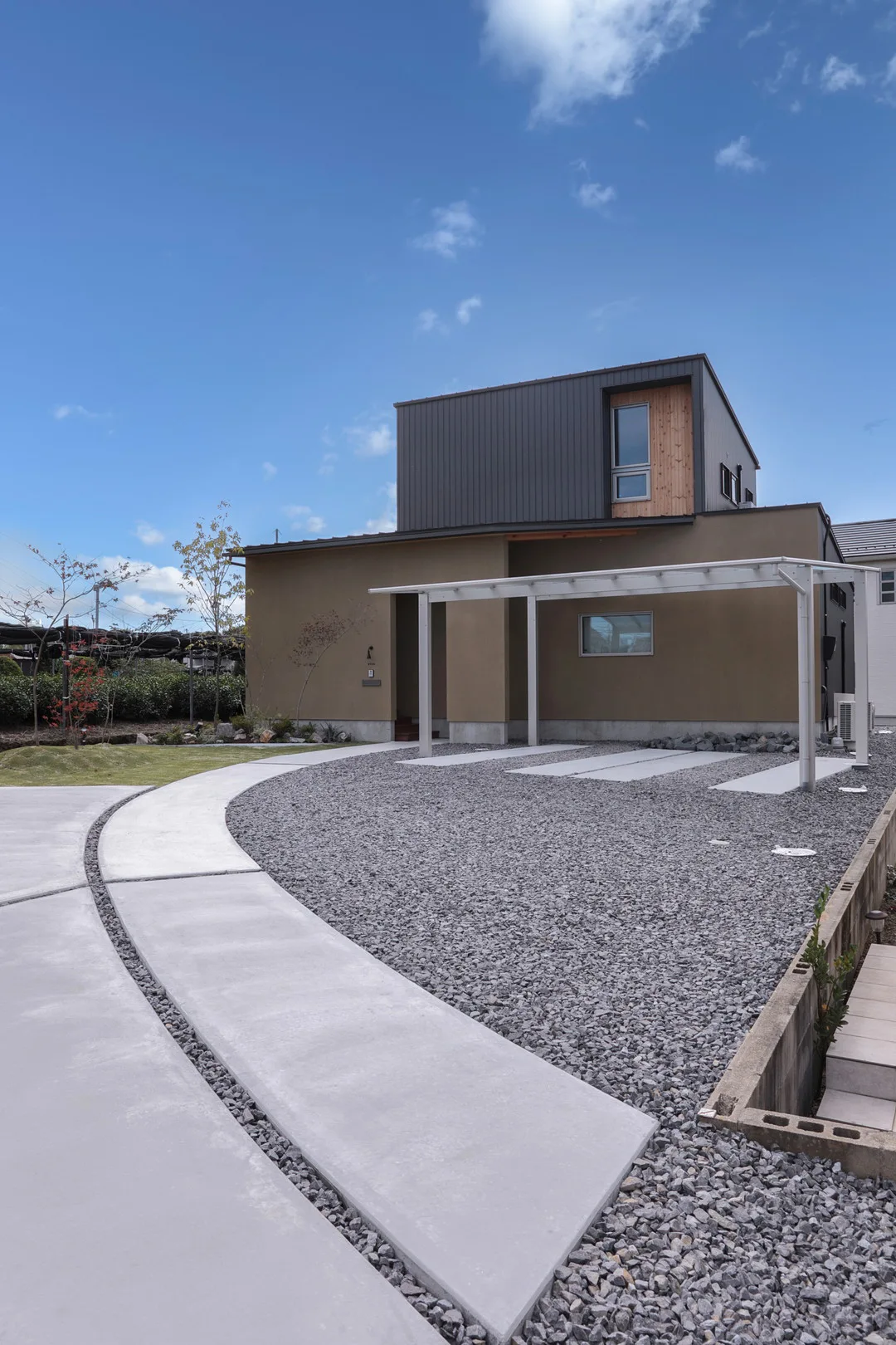 快晴の空の下に建つモダンな住宅の全景。ベージュと黒の外壁、手前にはカーポートと曲線を描くコンクリートのアプローチがある