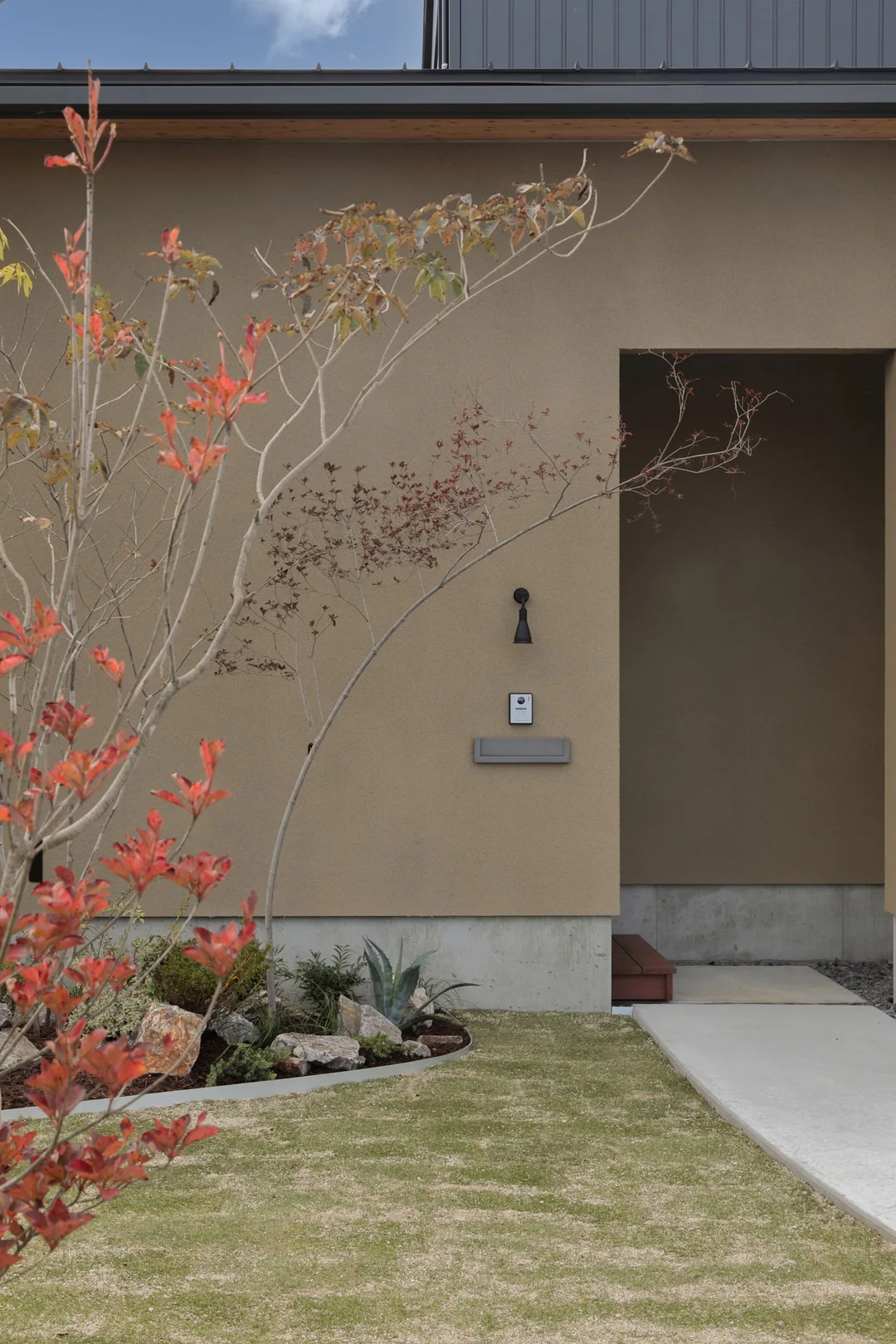 ベージュ色の外壁を持つ住宅の玄関アプローチ。黒い外灯、インターホン、ポストが設置され、手前には紅葉した木や植栽がある