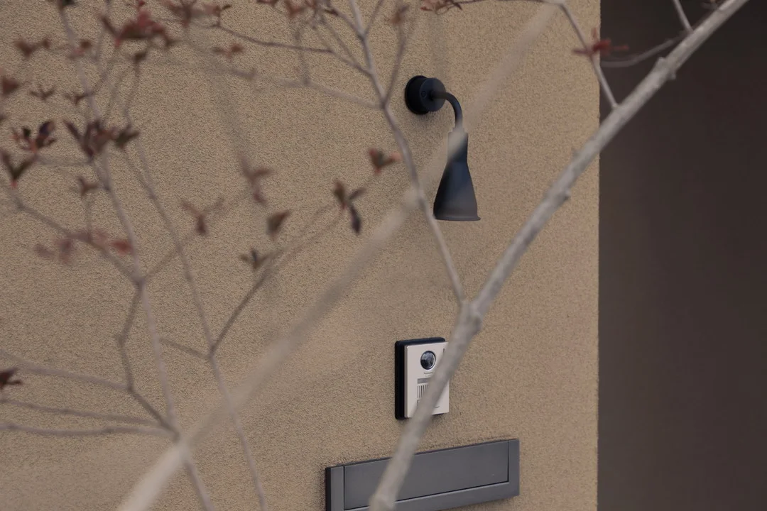 外壁に取り付けられた黒いブラケットライト、カメラ付きインターホン、ポストのクローズアップ。手前に細い木の枝が重なっている
